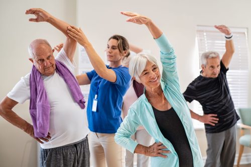 Active seniors doing exercise with physiotherapist at nursing home gym. Trainer helping elderly man and old woman exercising at home. Retired people doing stretching exercises at retirement community.